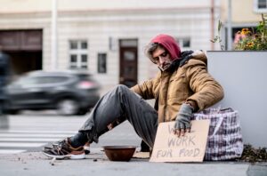 Homeless beggar man sitting outdoors in city asking for money donation.
